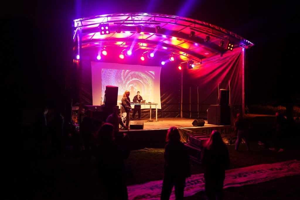 Ein DJ auf einer beleuchteten Bühne macht Musik für die anwesenden Personen.