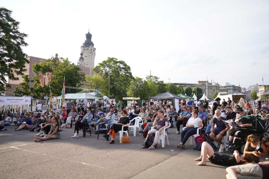 Menschen sitzen auf dem Wilhelm-Leuschner-Platz und sehen einem Bühnenprogramm zu.