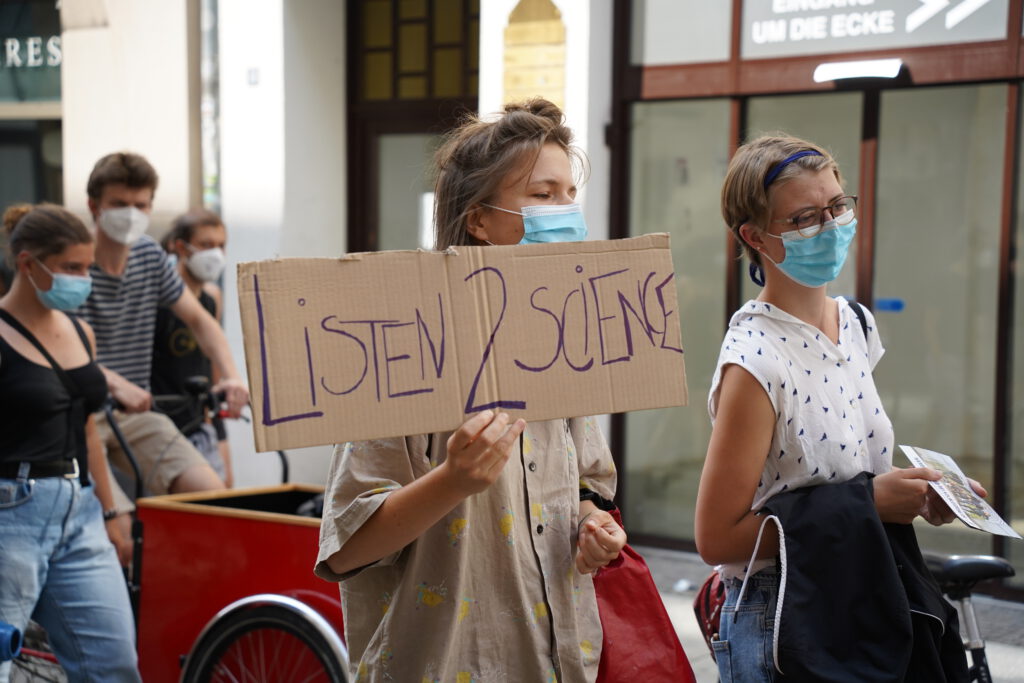 Zwei Personen laufen auf einer Demonstration. Die linke Person hält ein Schild, auf dem "Listen to science" steht.