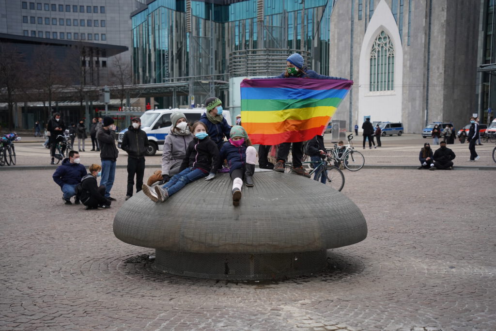 Einige Kinder sitzen auf dem Brunnen auf dem Augustusplatz. Eines der Kinder hält eine Regenbogen-Fahne hoch.