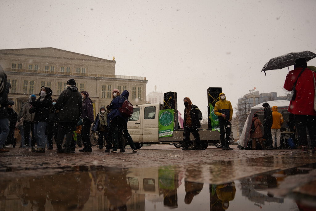 Menschen stehen auf dem Augustusplatz im Schneeregen, sie spiegeln sich in einer großen Pfütze.